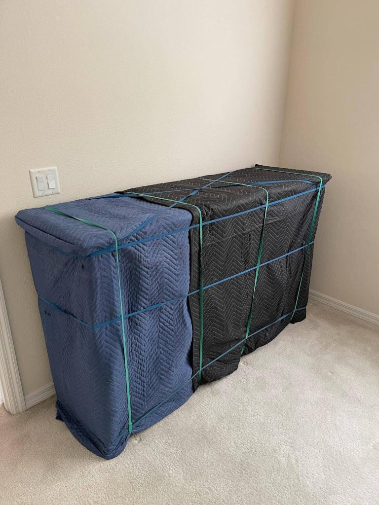Two dressers fully wrapped in blue and black moving pads and banded, standing against a wall in an empty room