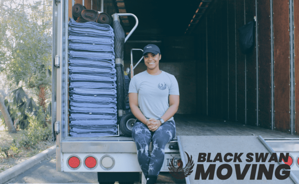 A smiling woman relaxes on the back of a moving truck, surrounded by neatly folded moving pads. Black Swan Moving logo visible on the side of the truck.