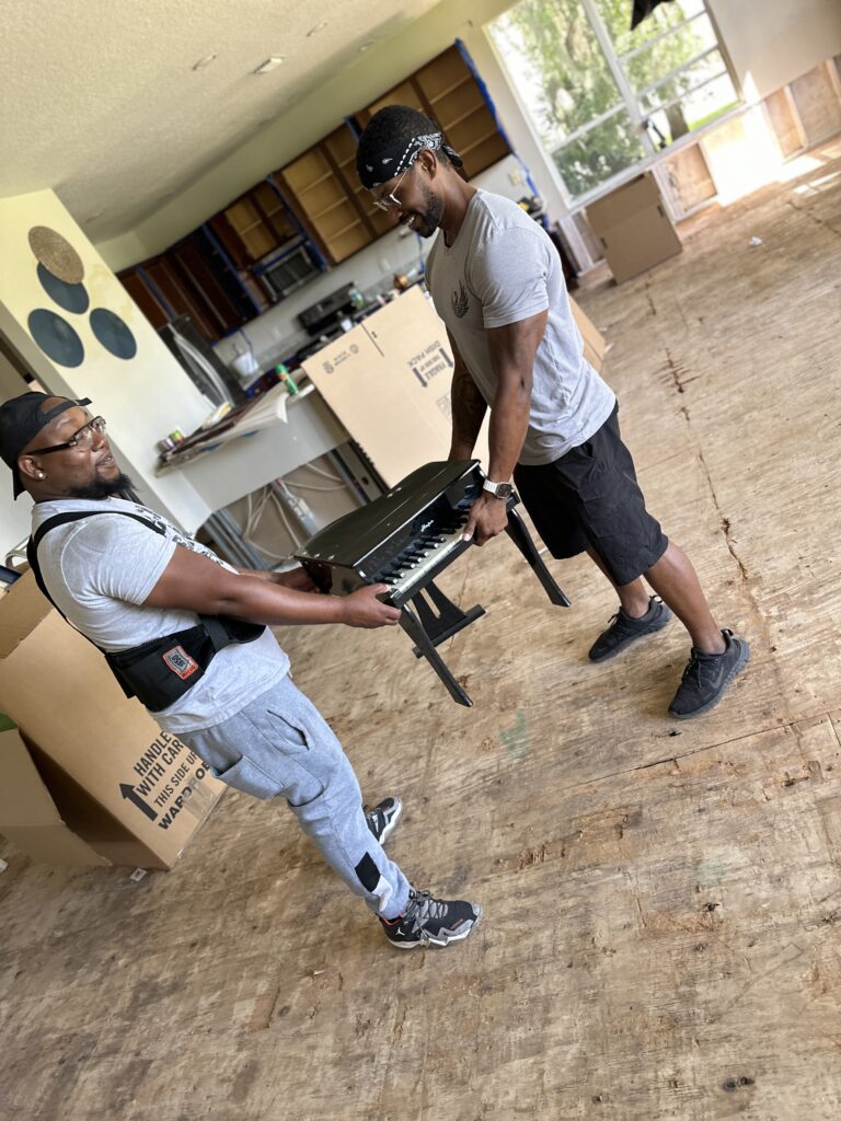 Two Black Swan movers jokingly carrying a child's toy grand piano