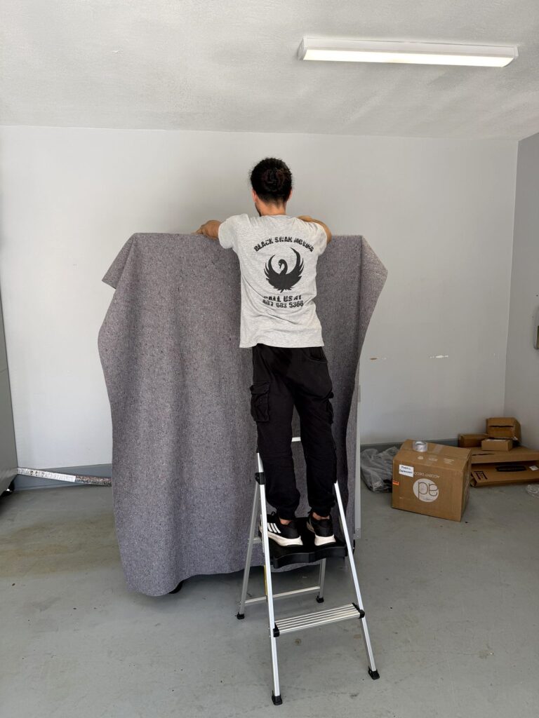 Tavares Movers - Black Swan Moving | 5-Star Movers in Central Florida Black Swan Moving crew member on a step ladder draping a gray moving pad over a tall piece of furniture