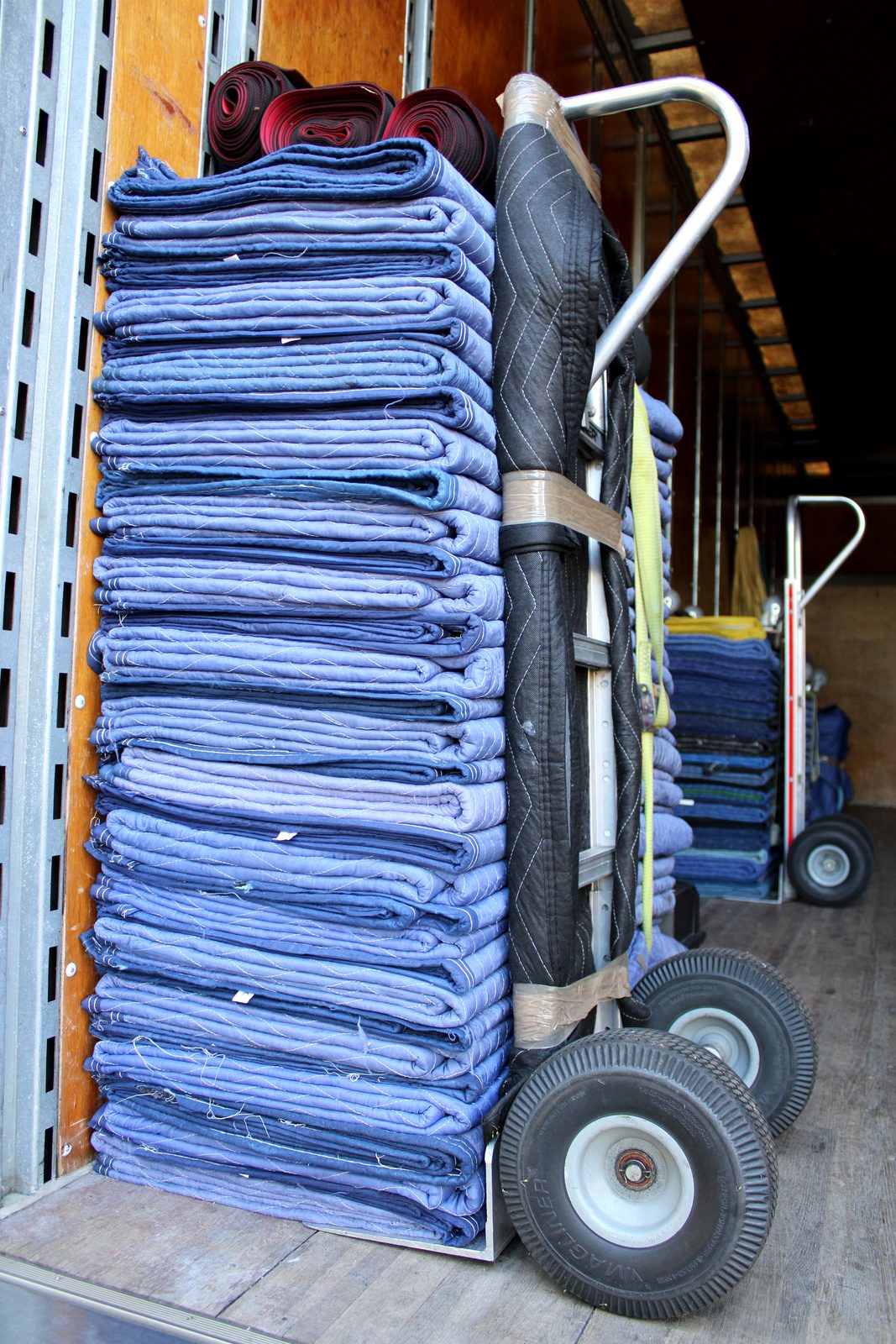 Hand truck stacked with clean folded blue moving pads beside the Black Swan Moving truck