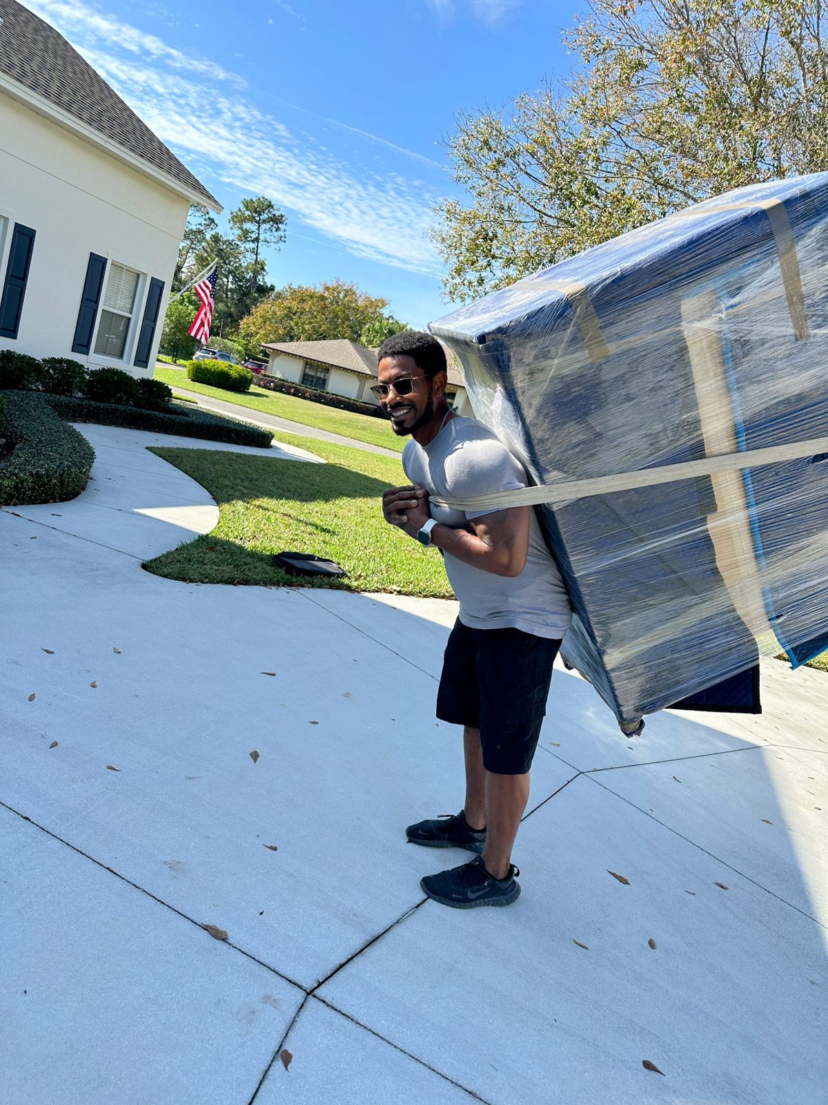 Black Swan Moving mover carrying a blanket-padded, shrink-wrapped piece of furniture on his back with a carrying strap across a driveway