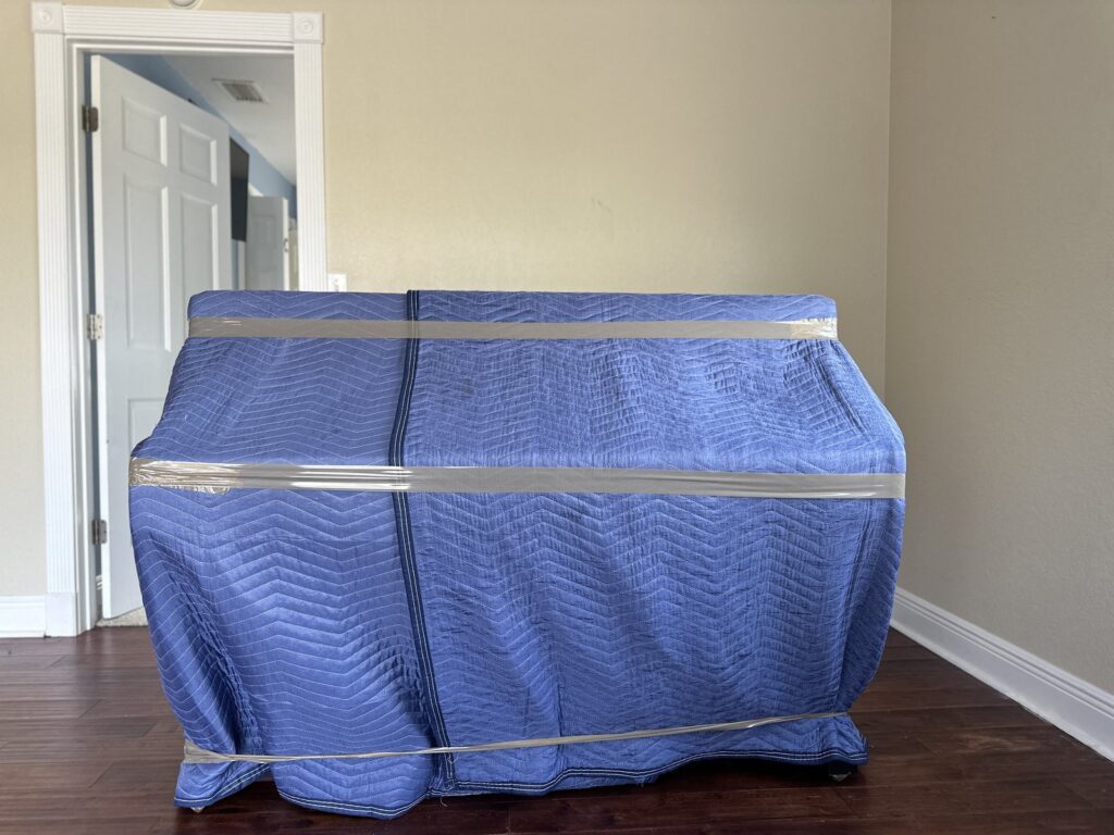 Upright piano wrapped in blue moving blankets and strapped, ready to move