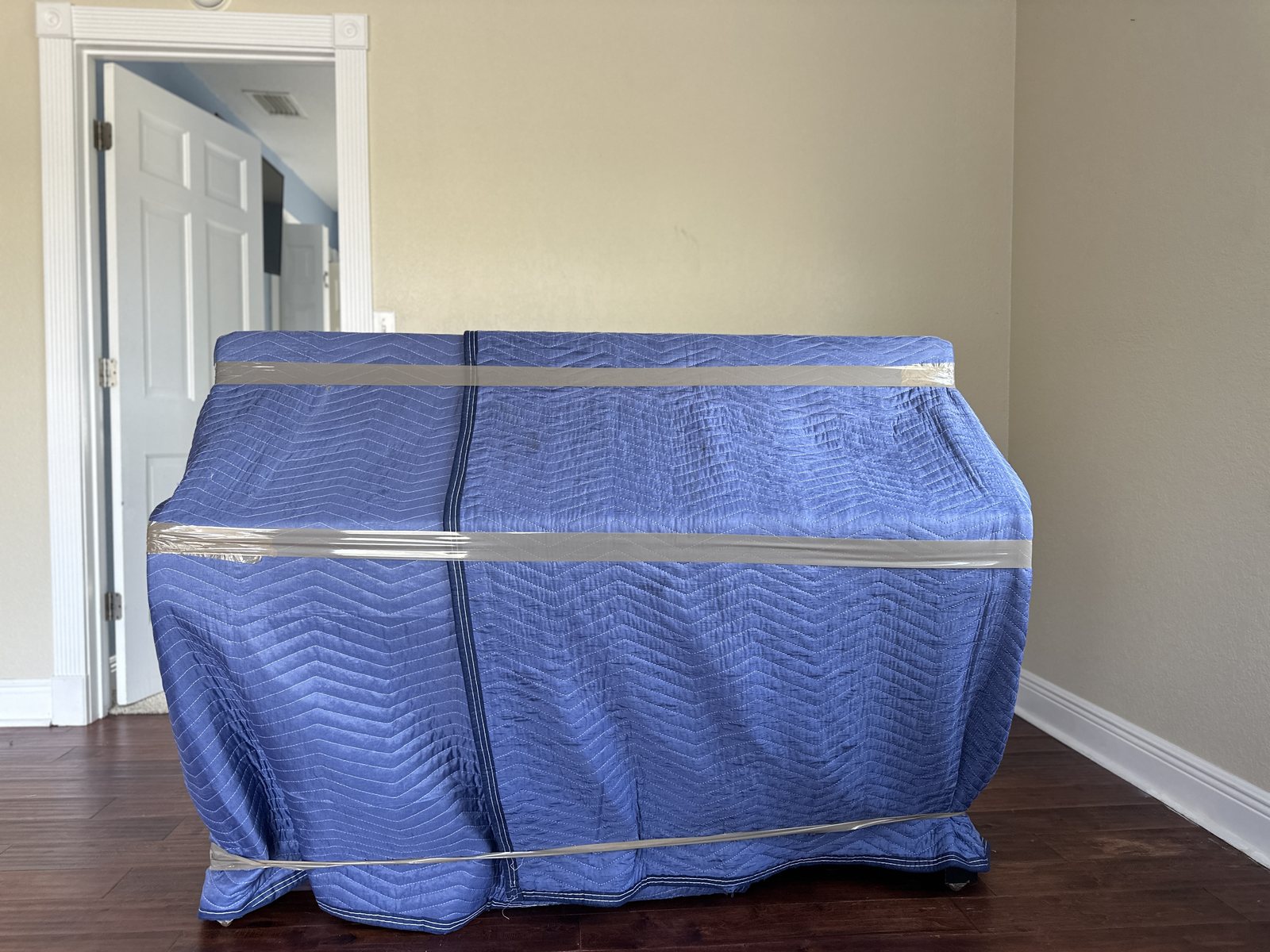 Upright piano fully covered in blue moving blankets held tight with tape bands, standing on a hardwood floor before the move