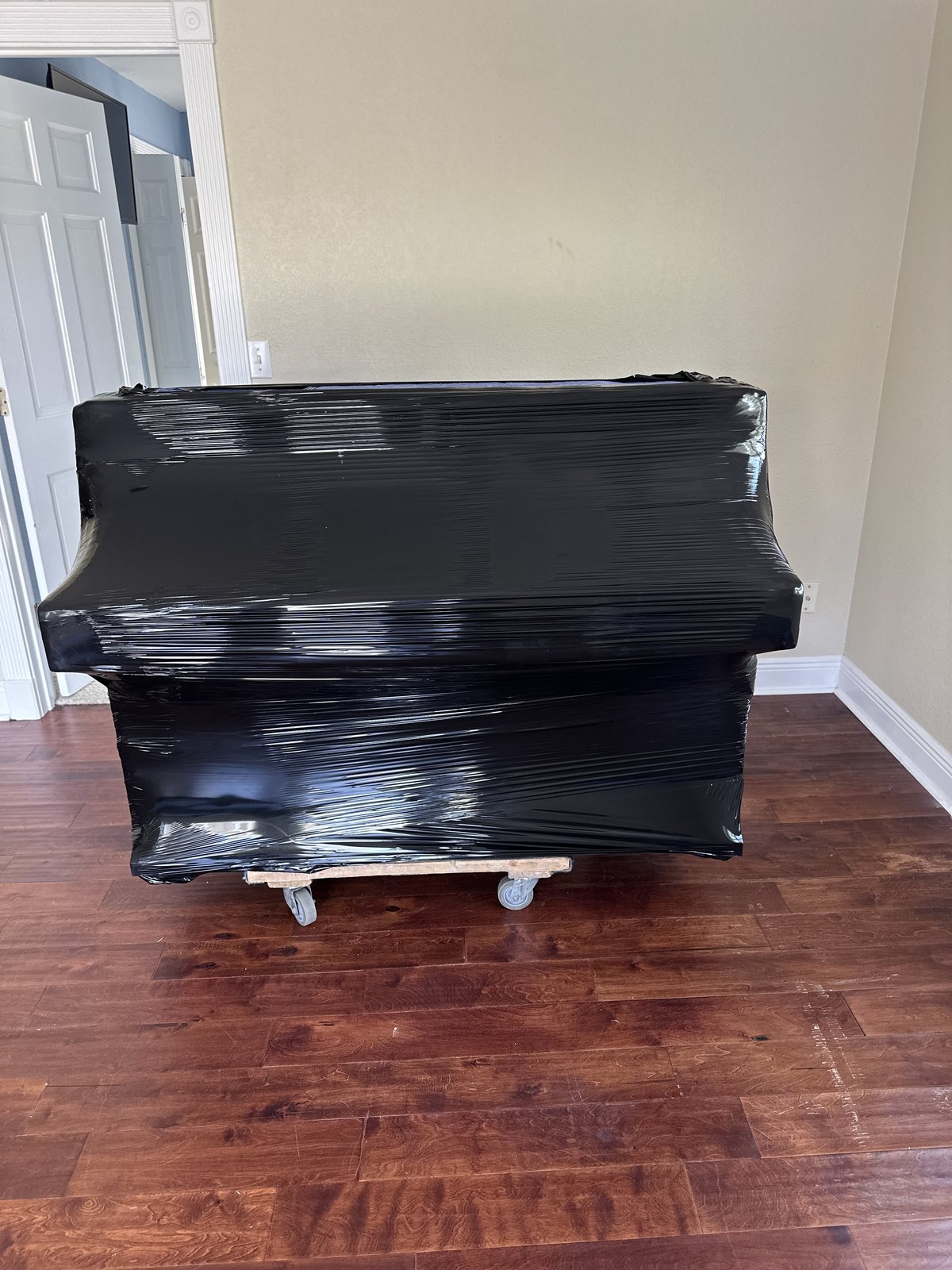 Upright piano shrink-wrapped in black plastic over moving blankets, resting on a wooden piano dolly with wheels