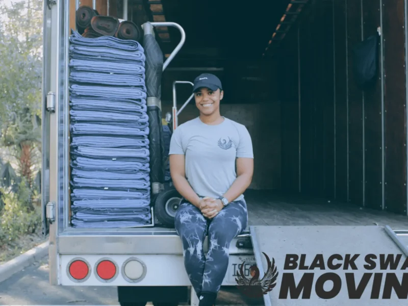 A smiling woman relaxes on the back of a moving truck, surrounded by neatly folded moving pads. Black Swan Moving logo visible on the side of the truck.
